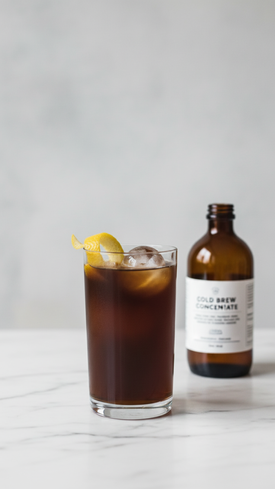 Cold brew and tonic with large clear ice sphere and wide lemon peel twist on white marble countertop
